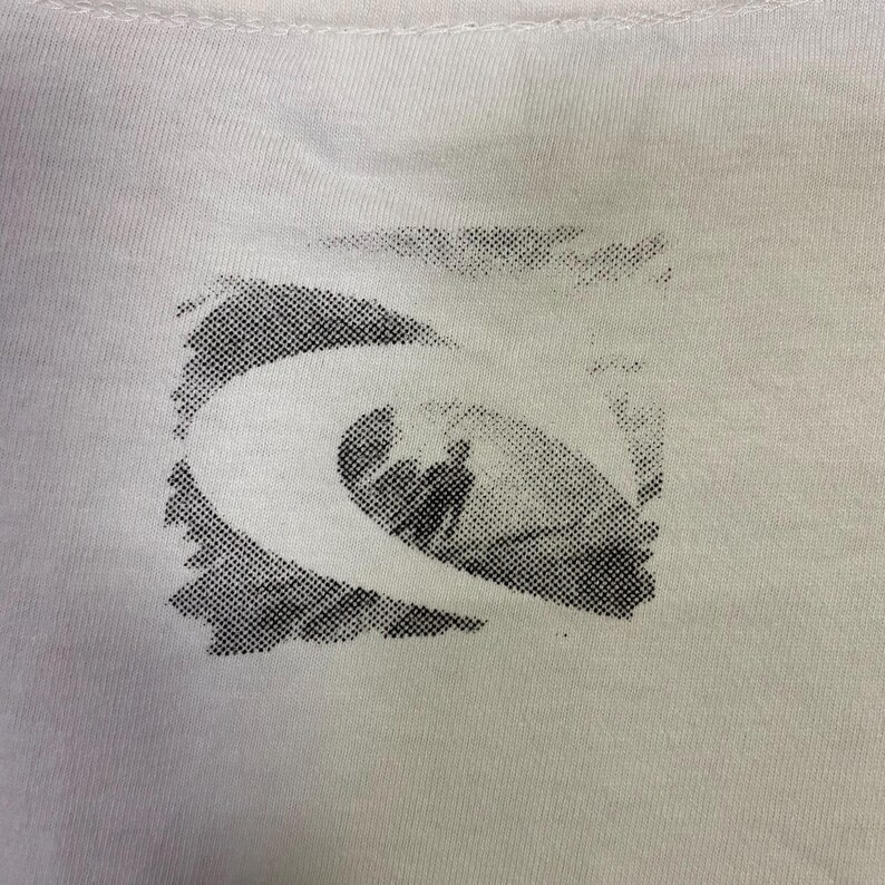 May include: Close-up of a white t-shirt featuring a black and white graphic design. The design is a stylized wave with a surfer silhouette inside a square frame. The shirt has a crew neck.