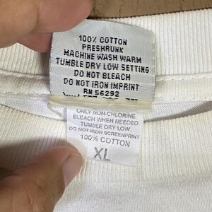 May include: Close-up of a white fabric tag with black text. The tag includes washing instructions: machine wash warm, tumble dry low, and do not bleach. The tag also indicates the fabric is 100% cotton and the size is XL.