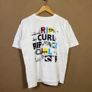 May include: White t-shirt with the text "RIP CURL" in a colorful, layered design. The text is in red, yellow, blue, green, and black. The shirt is hanging on a wooden hanger.
