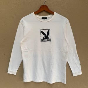May include: A white long-sleeved shirt featuring the black Playboy logo. The logo is a black bunny head within a black square, with the word "PLAYBOY" beneath. The shirt is displayed on a wooden hanger.