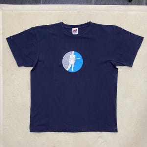 Puede incluir: Una camiseta azul marino con una silueta blanca de una persona tocando la guitarra dentro de un círculo azul y blanco. El logotipo de New Balance es visible en la camiseta.