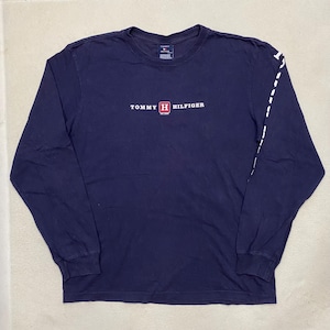 May include: A navy blue long-sleeve t-shirt with a white Tommy Hilfiger logo and a red box with a white "H" inside. The shirt has white stitching on the sleeves.