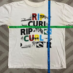May include: White t-shirt with the text "RIP CURL" in a colorful, layered design. The shirt features a crew neck and short sleeves. The shirt measures approximately 24 inches across the chest.