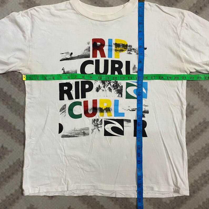 May include: White t-shirt with the text "RIP CURL" in a colorful, layered design. The shirt features a crew neck and short sleeves. The shirt measures approximately 24 inches across the chest.