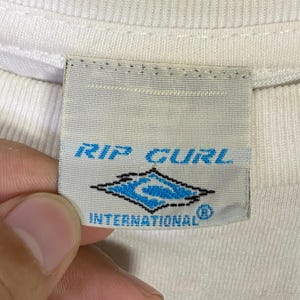 May include: Close-up of a white fabric tag with the text "RIP CURL" in blue. Below the text is a blue and white diamond-shaped logo. The text "INTERNATIONAL" is below the logo.