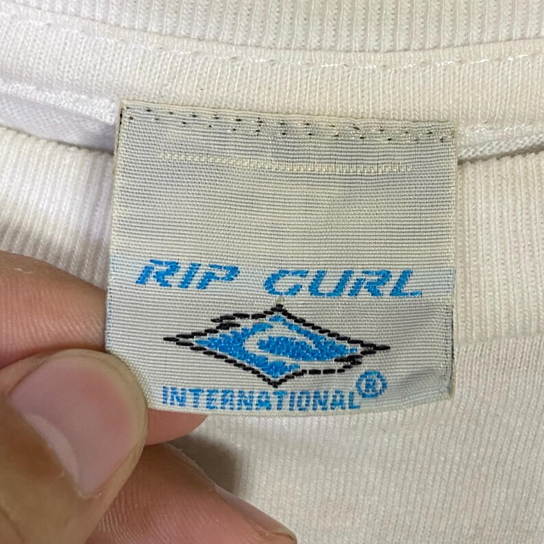May include: Close-up of a white fabric tag with the text "RIP CURL" in blue. Below the text is a blue and white diamond-shaped logo. The text "INTERNATIONAL" is below the logo.