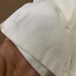 May include: Close-up of a white fabric, possibly a t-shirt or other garment. The edge of the fabric is visible, showing a neatly sewn seam with a series of small stitches. The fabric appears to be soft and slightly textured.