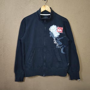 May include: Dark navy zip-up sweatshirt with a high collar and two front pockets. The sweatshirt has a graphic design on the front, including the brand logo in red and white. The garment is hanging on a wooden hanger.