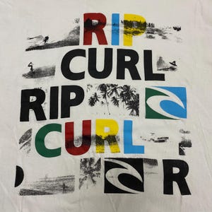 May include: White t-shirt with the words "RIP CURL" in large, colorful letters. The design includes images of surfers, palm trees, and the Rip Curl logo. The letters are in red, yellow, blue, and green.