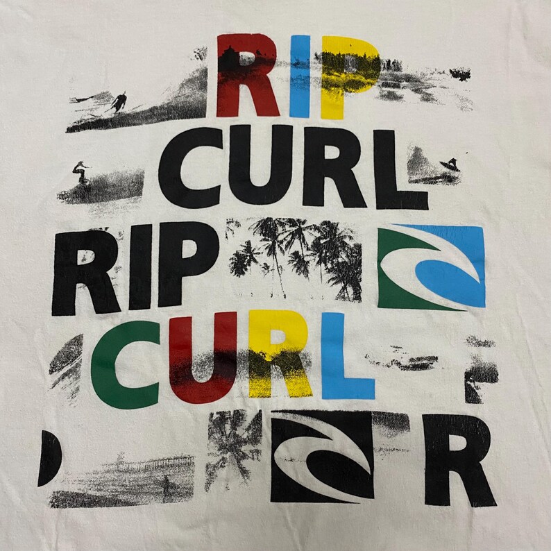 May include: White t-shirt with the words "RIP CURL" in large, colorful letters. The design includes images of surfers, palm trees, and the Rip Curl logo. The letters are in red, yellow, blue, and green.