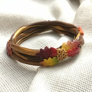 May include: A brown and gold clay bracelet with a twisted design. The bracelet is decorated with fall-colored leaves and berries.