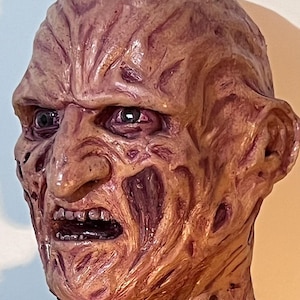 May include: A realistic mask depicting a character with a severely scarred, textured, and discoloured face in shades of tan, brown, and deep red. The mask features a prominent nose, deep-set eyes, and an open mouth revealing teeth.