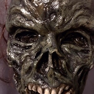 May include: A close-up of a detailed, green and grey zombie mask with a wide open mouth and visible teeth. The mask is made of a textured material that gives it a realistic look.