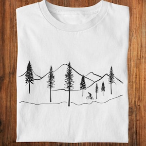 May include: White t-shirt featuring a black graphic of a mountain range, pine trees, and a cyclist. The design is minimalist and outdoors-themed, perfect for those who enjoy biking and nature. The shirt is folded on a wooden surface.