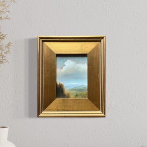 May include: A small framed painting of a landscape with a blue sky, white clouds, and green hills. The frame is gold and has a simple design.