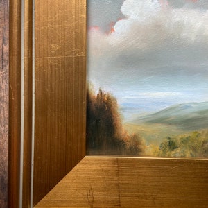 May include: A framed oil painting depicting a landscape scene with a cloudy sky, rolling hills, and a distant body of water. The frame is a gold color with a double border.