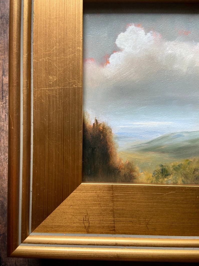 May include: A framed oil painting depicting a landscape scene with a cloudy sky, rolling hills, and a distant body of water. The frame is a gold color with a double border.