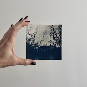 May include: A square, cyanotype print featuring a silhouette of a house and foliage against a light background. The dark blue image is held by a hand with dark nail polish. The print evokes a sense of nature and architecture.
