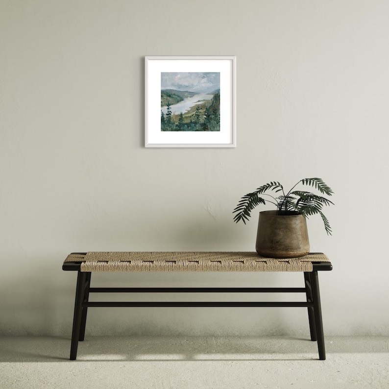 May include: A framed painting of a landscape with a river winding through a valley. The painting is in a white frame and is hanging on a light gray wall.