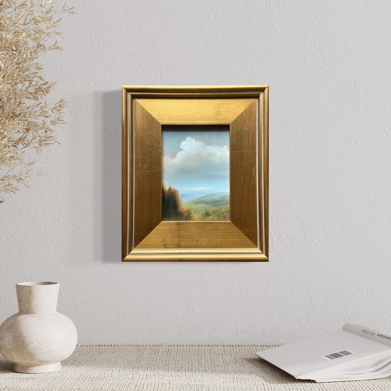 May include: A small framed painting of a landscape with a blue sky, white clouds, and green hills. The frame is gold and has a simple design.