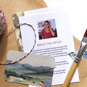 May include: A close-up of a card with a watercolor painting of a landscape. The card is on a brown surface with a roll of twine, a paintbrush, and a piece of washi tape. The text on the card reads "About the Artist".