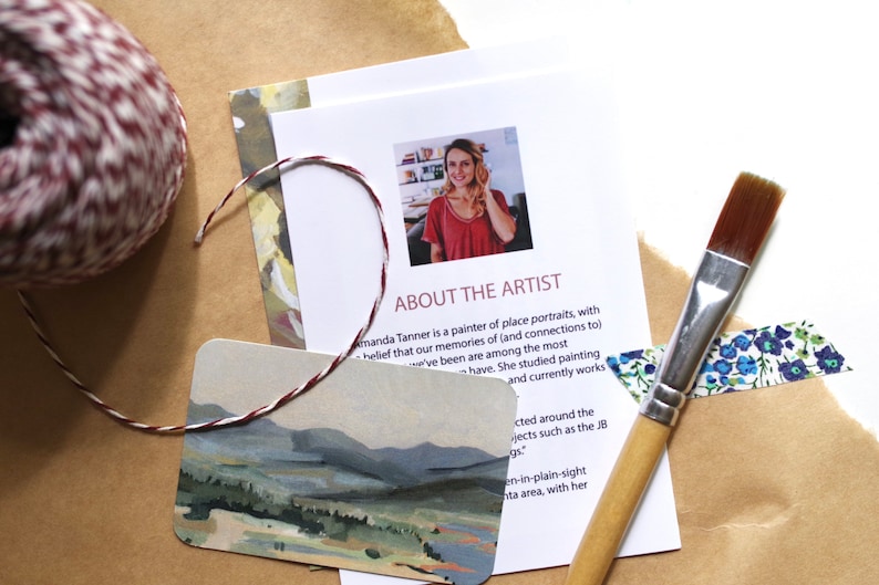 May include: A close-up of a card with a watercolor painting of a landscape. The card is on a brown surface with a roll of twine, a paintbrush, and a piece of washi tape. The text on the card reads "About the Artist".