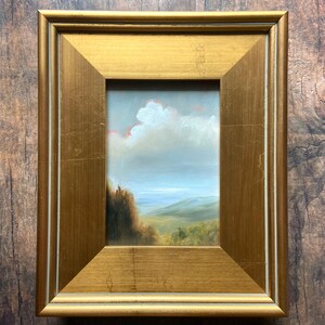 May include: A framed oil painting of a landscape with a cloudy sky, green hills, and a body of water in the distance.