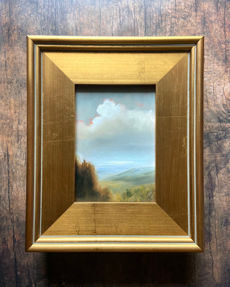 May include: A framed oil painting of a landscape with a cloudy sky, green hills, and a body of water in the distance.
