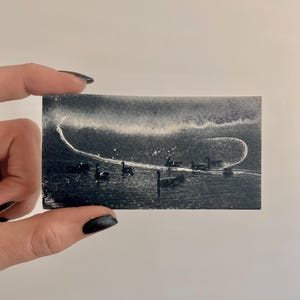 May include: A small, rectangular, black and white painting depicting a body of water with several dark silhouettes of birds. A white, curved line arcs across the top of the painting, and the image is held by a hand.