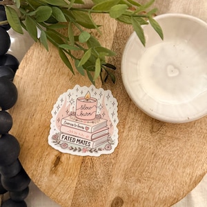 May include: A white ceramic mug with a black handle and the text "But First, Coffee" in a bold, black font. The mug is set against a blurred background, suggesting a cozy setting.