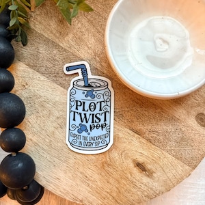 May include: A sticker in the shape of a soda can with the words "PLOT TWIST pop" and "EXPECT THE UNEXPECTED IN EVERY SIP" in a whimsical font. The can is light blue with bubbles and a blue straw.