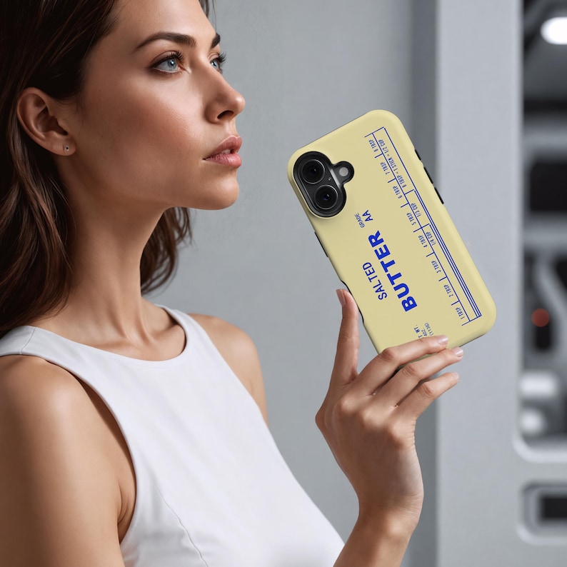 May include: A phone case with a butter-themed design. The case is light yellow with blue text that reads "SALTED BUTTER AA GRADE". The design includes measurement markings along the top edge. The phone case is held by a person.