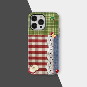 Puede incluir: Una funda de teléfono patchwork con una parte superior a cuadros verdes, un centro de cuadros rojos y blancos y un lado de mezclilla. Adornos incluyen un borde de encaje blanco, una manzana roja, una cereza roja y una oruga verde.