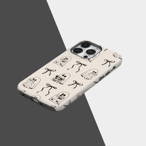 May include: A cream-colored phone case featuring a repeating pattern of black and white ghost illustrations and black bows. The case has a glossy finish and is designed to fit a smartphone, with cutouts for the camera and buttons.