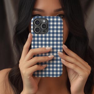 May include: A navy blue and white gingham pattern phone case. The case has a square grid design and a camera cutout. The phone case is held up in front of a person's face.