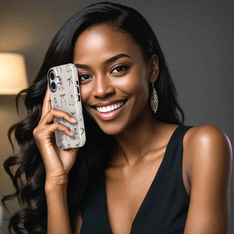 May include: A smartphone case with a beige background and a repeating pattern of black ghost and bow illustrations. The case is held up in front of a person.