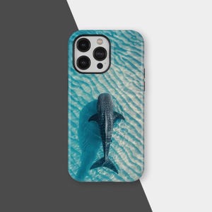 May include: A phone case depicting a whale shark in turquoise water. The case has a blue border and a glossy finish. The whale shark is dark with white spots, and the water has a rippled sand pattern. The case is designed for a smartphone.