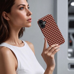 May include: A smartphone case with a red and beige gingham pattern. The case has a black border and a camera cutout. The phone case is held by a person wearing a white top.