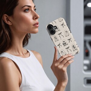 May include: A beige phone case with a repeating pattern of black line-drawn ghosts and bows. The case has a glossy finish and a camera cutout. The design is simple and whimsical, perfect for Halloween or everyday use.