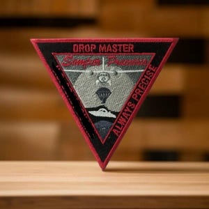 May include: A red, black, and gray triangular patch with the text "DROP MASTER" and "ALWAYS PRECISE" in red. The patch features a stylized image of a parachute and a plane.