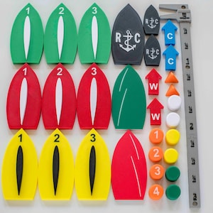 Op de afbeelding: Een verzameling kleurrijke navigatiehulpmiddelen voor de watersport, waaronder groene, rode en gele boeien met nummers, pijlen en markeringen. Bevat ook grijze markeringen met de letters "R C" en een ankersymbool, en een grijze liniaal.