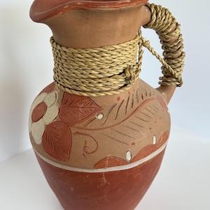 May include: Earthenware pitcher with a reddish-brown base and rim, adorned with a floral and leaf design in shades of red and white. The handle is wrapped with woven straw, adding a rustic touch. A decorative piece for the home.
