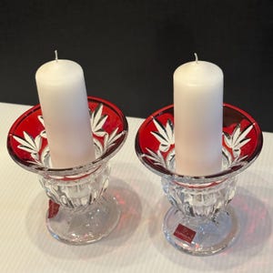 May include: Two clear glass candle holders with red and white accents, each holding a white pillar candle. The candle holders have a decorative design and a round base. The red accents feature a leaf-like pattern.