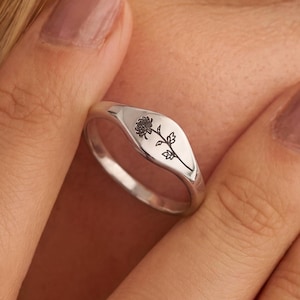 May include: A silver signet ring featuring a delicate, black engraved floral design on its flat, oval-shaped face. The ring has a polished, smooth metallic finish and a slender band, showcasing a detailed botanical illustration of a single flower.
