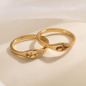 May include: Two gold-toned metal rings featuring delicate, thin bands and flat, circular face plates. Each ring is engraved with a small, detailed black botanical illustration of a single flower stem. The rings have a polished, reflective finish.