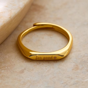 Peut inclure: Une bague dorée avec l'inscription "for good" en écriture fine. La bague a une face rectangulaire et une finition lisse et polie. La bague est posée sur un fond neutre. Bijou élégant et simple.