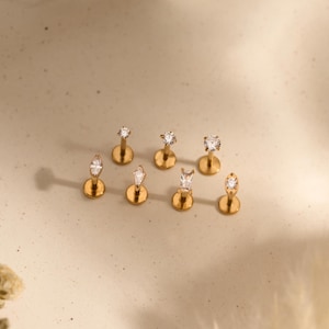 May include: A collection of seven gold-tone flat back stud earrings, each featuring a single clear gemstone in various shapes including round, marquise, and baguette cuts. The jewelry pieces are arranged in a cluster, and the text Jupiter Bloom is visible at the bottom.