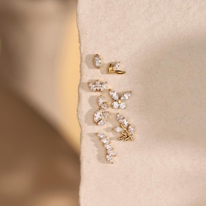 May include: A collection of gold-toned earrings with sparkling clear gemstones. The earrings feature various designs, including a butterfly, a floral sprig, and a curved bar. The image is set against a neutral background, with the text "Jupiter Bloom" at the bottom.