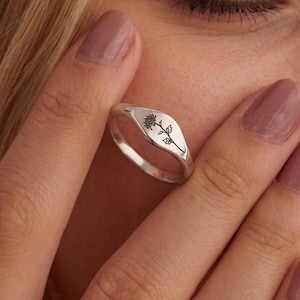May include: A silver ring with a floral design engraved on the oval face. The ring is held up to a person's face, with the Jupiter Bloom logo visible at the bottom.