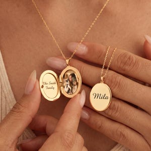 May include: A gold oval locket necklace on a delicate chain. The locket is open, revealing a small photograph and the text The Smith Family. A second closed gold oval locket hangs nearby, engraved with the name Mila in a cursive script.